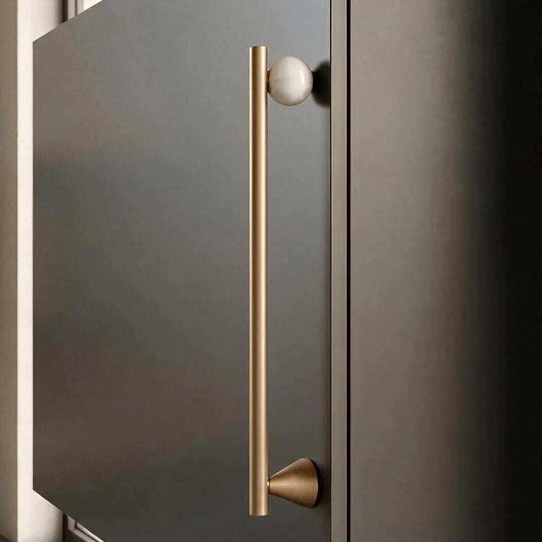 A modern long bar cabinet handle in brushed brass with a spherical white knob, mounted on a dark charcoal-gray door.