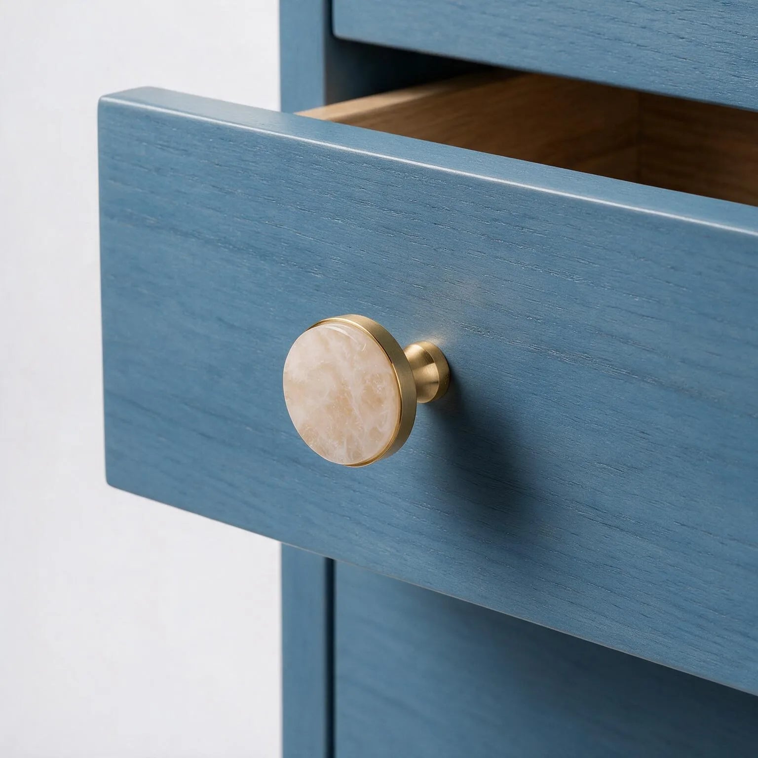 A close-up of a blue wooden drawer with an open top drawer showing a wood interior, featuring a round cream marble-look knob with a brass base.