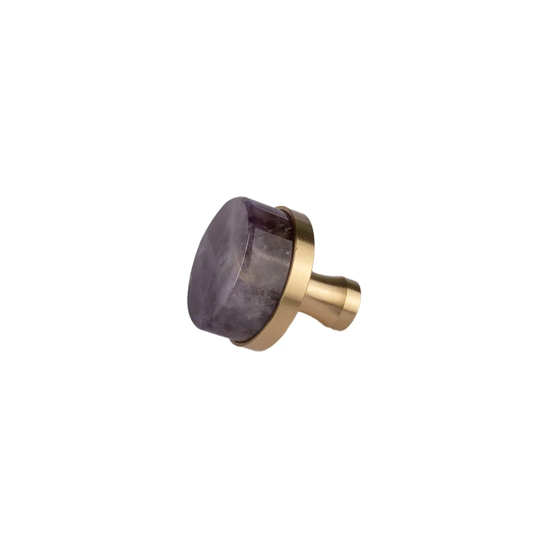 A cabinet knob featuring a polished, rounded purple amethyst stone inlay set within a brushed gold brass frame, shown against a solid white background.