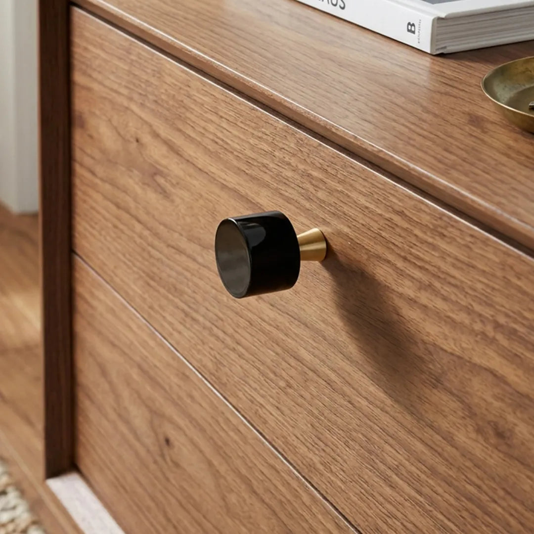 A close-up of a wooden dresser drawer featuring a modern cylindrical black knob with a brushed gold base, set against a warm wood grain background.