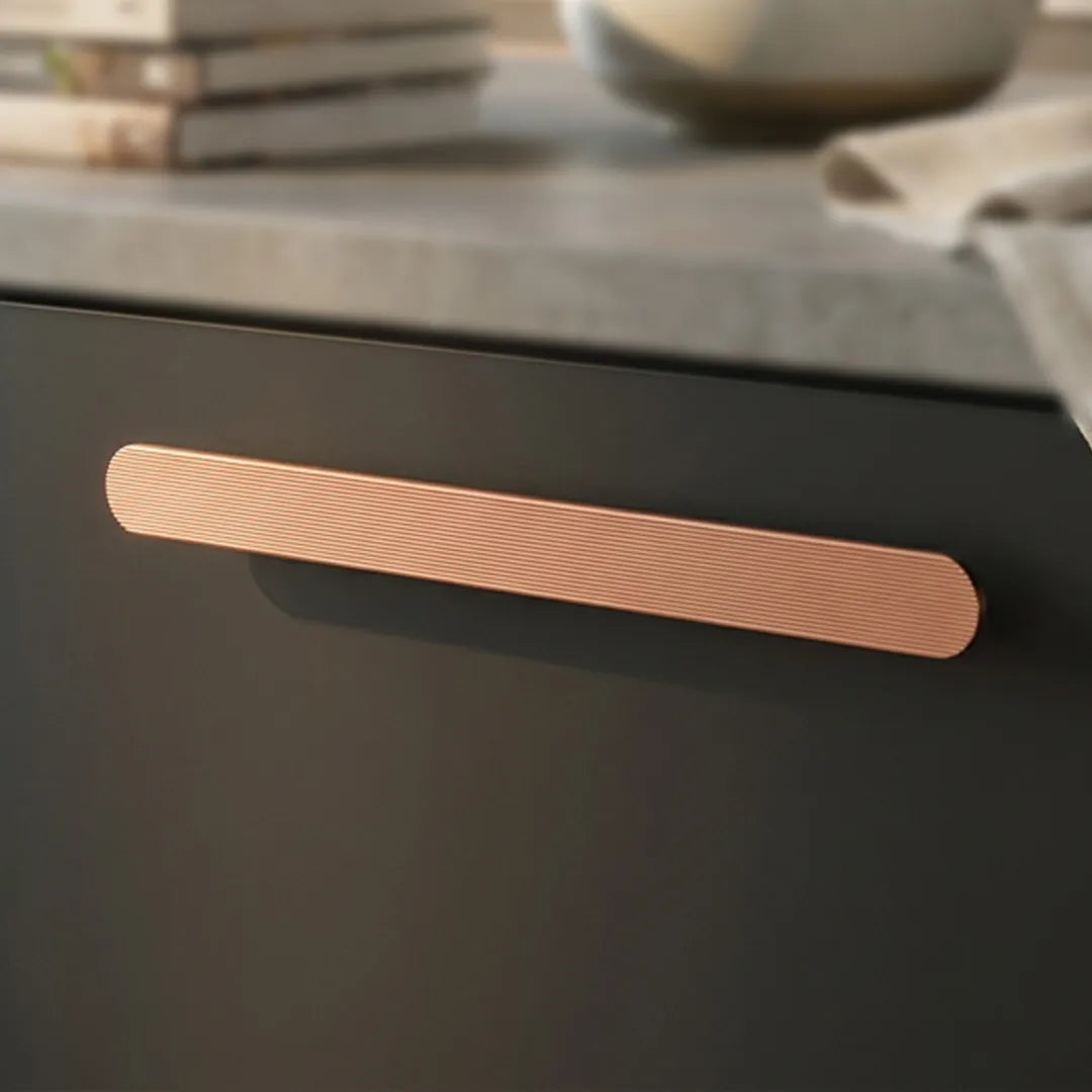 A modern elongated cabinet handle in a brushed rose gold finish mounted on a dark charcoal-gray door, with a blurred kitchen countertop and books in the background.