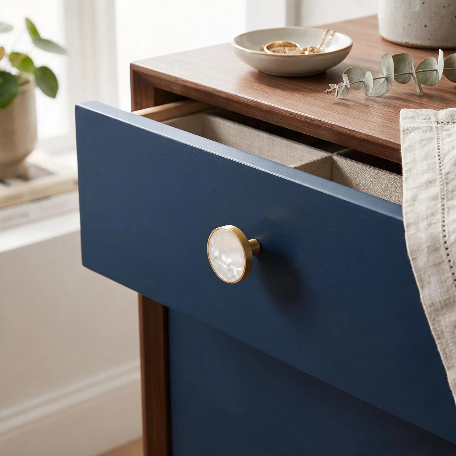 A close-up of a navy blue drawer on a wooden nightstand with a round brass knob, shown in a styled home setting with a bowl and eucalyptus leaves on top.