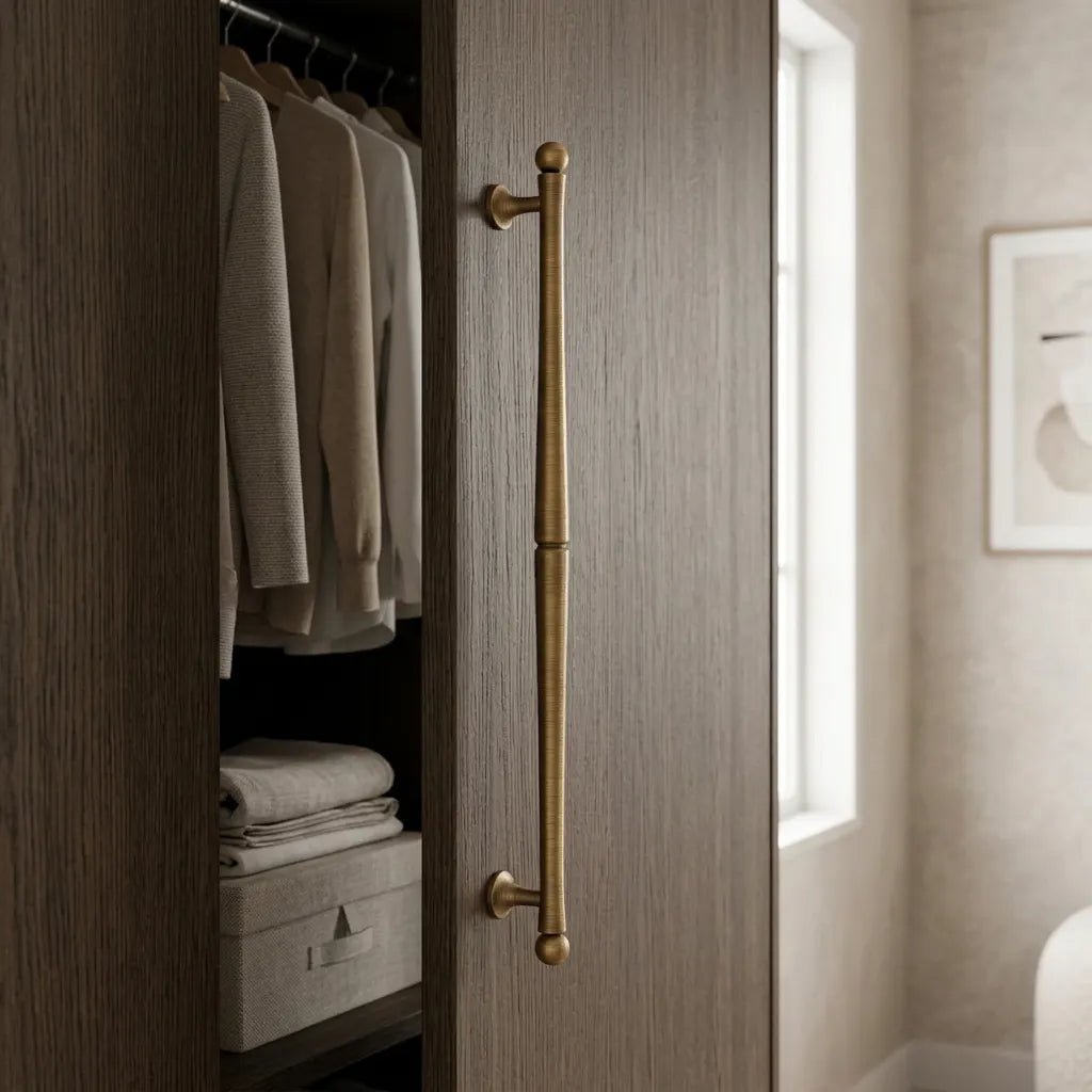 A tall, vertical brushed brass closet door handle installed on a wood-grain closet door, with folded sweaters and storage bins visible inside the open wardrobe in a softly lit room.