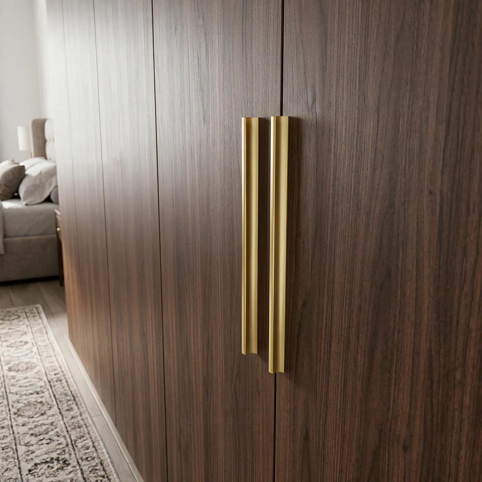 A pair of modern, slim vertical brass door handles mounted on dark wood grain doors in a bedroom setting.