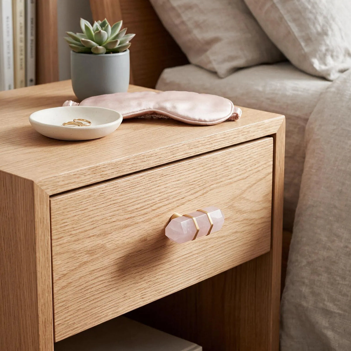 A light oak wood nightstand with a geometric crystal handle, styled in a bedroom next to a bed with beige linens. On top of the nightstand are a pink satin sleep mask, a small white dish with rings, and a potted succulent.