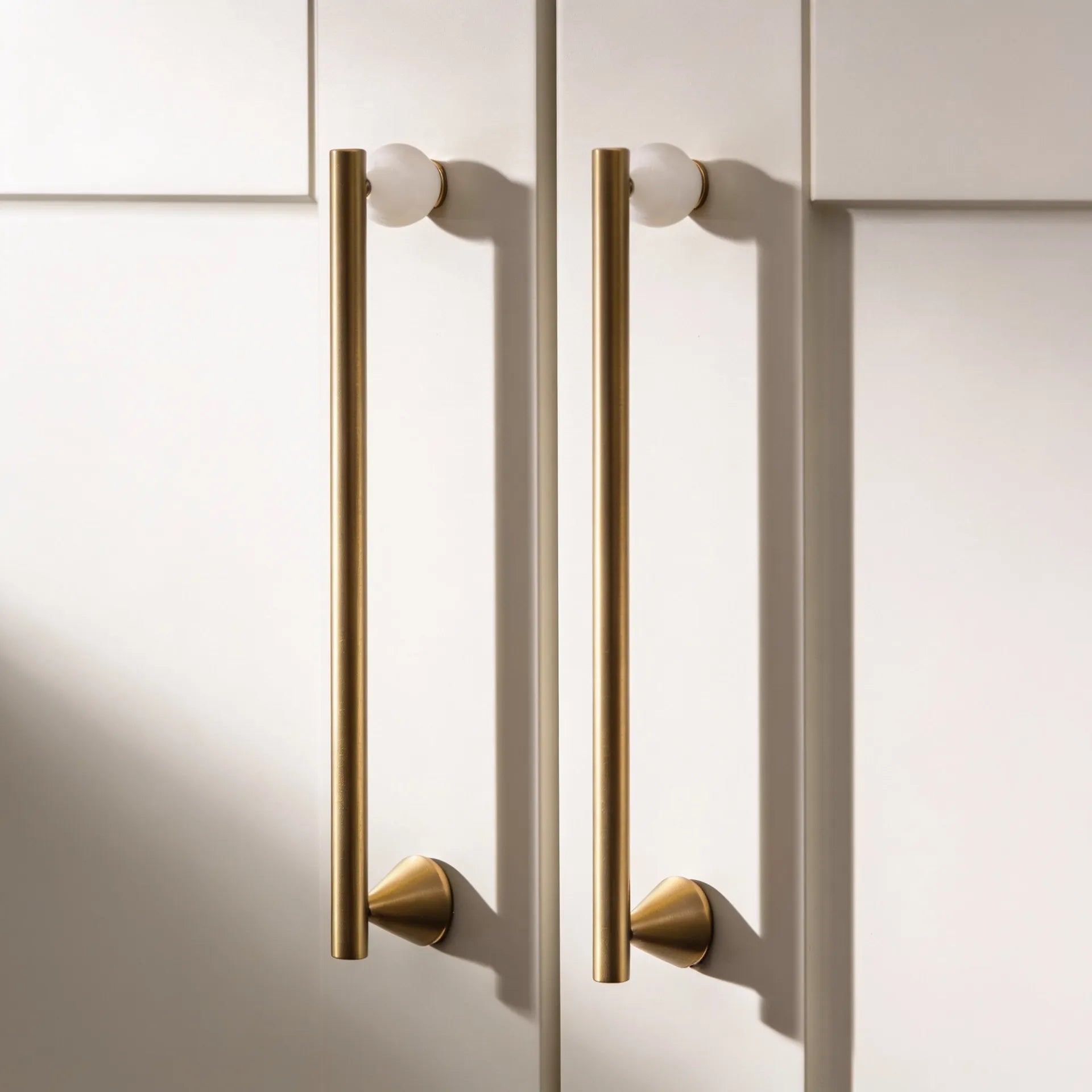Two long vertical brass cabinet handles with round white knobs, mounted on a cream-colored paneled cabinet door.