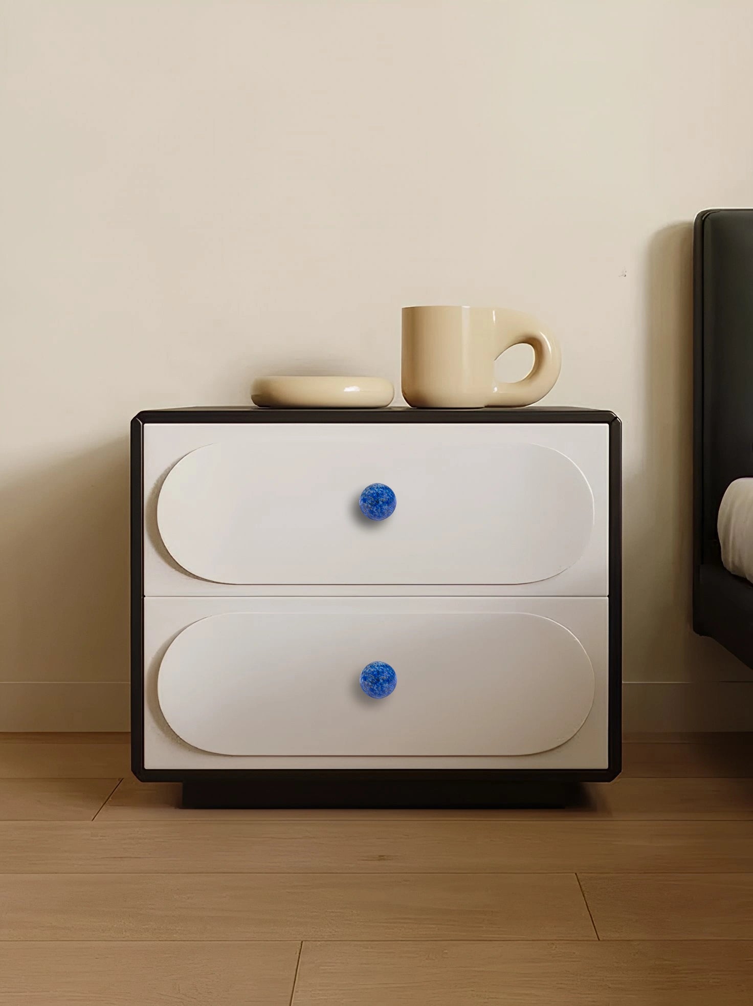 White nightstand with blue knobs, topped with a cup and saucer, placed on a wooden floor, styled by Mantara.