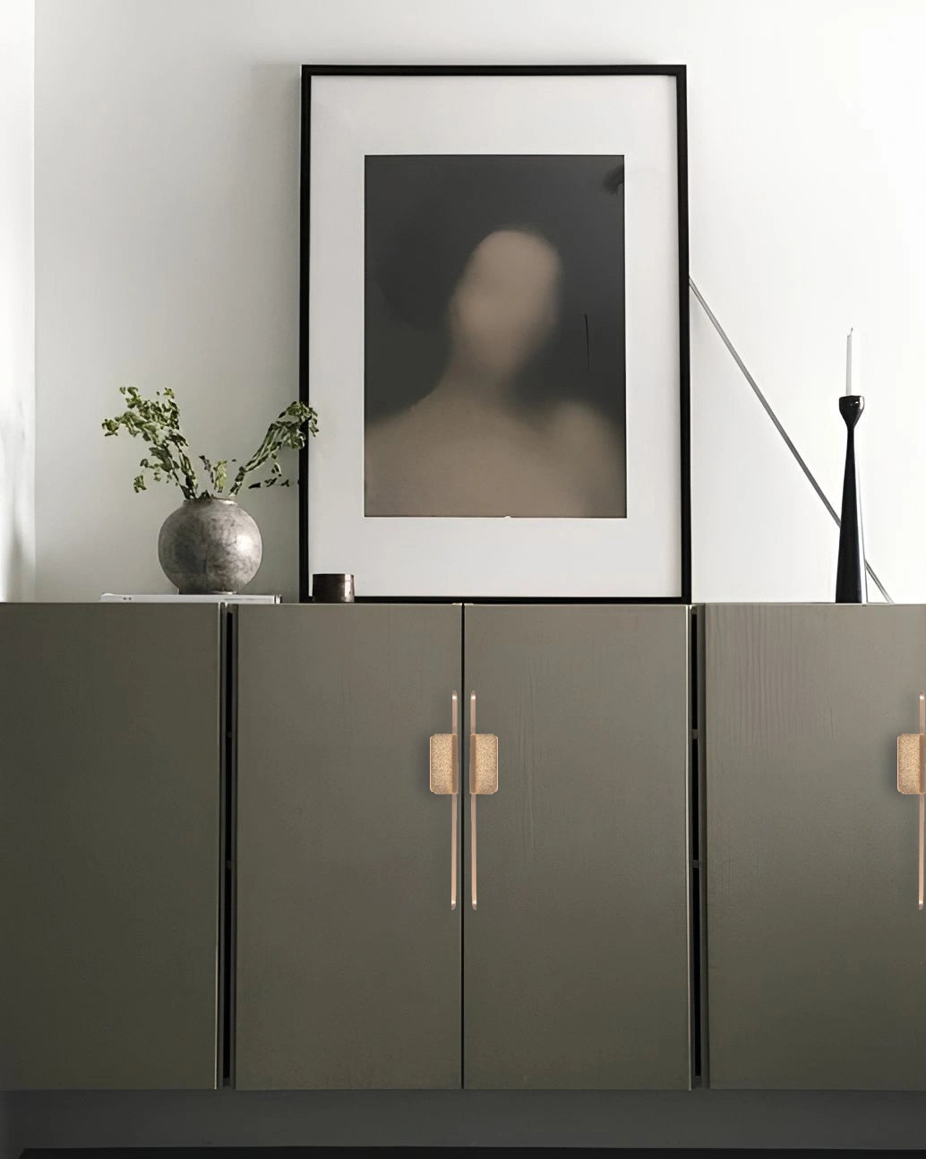Modern interior with a dark green cabinet featuring sleek handles, framed abstract art, and a vase with greenery—by Mantara.