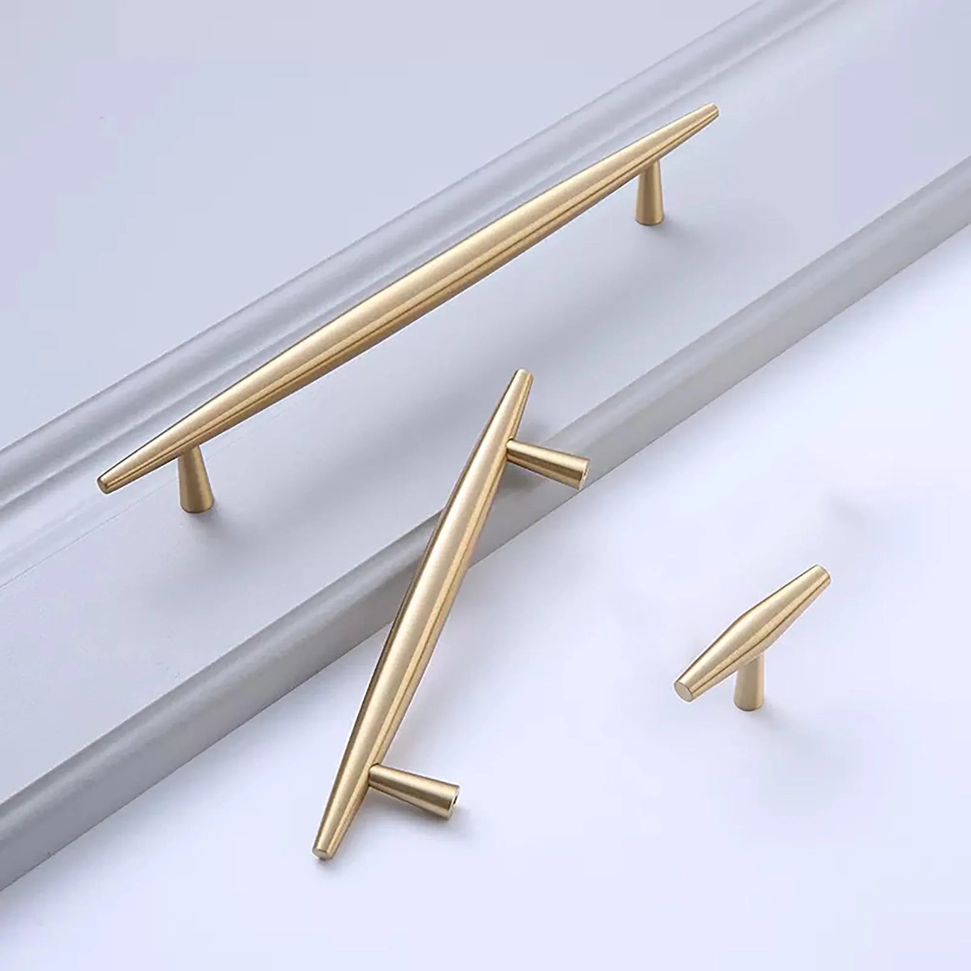 Three sleek, brass cabinet handles of varying lengths, showcasing modern design with tapered ends, placed on a light surface.