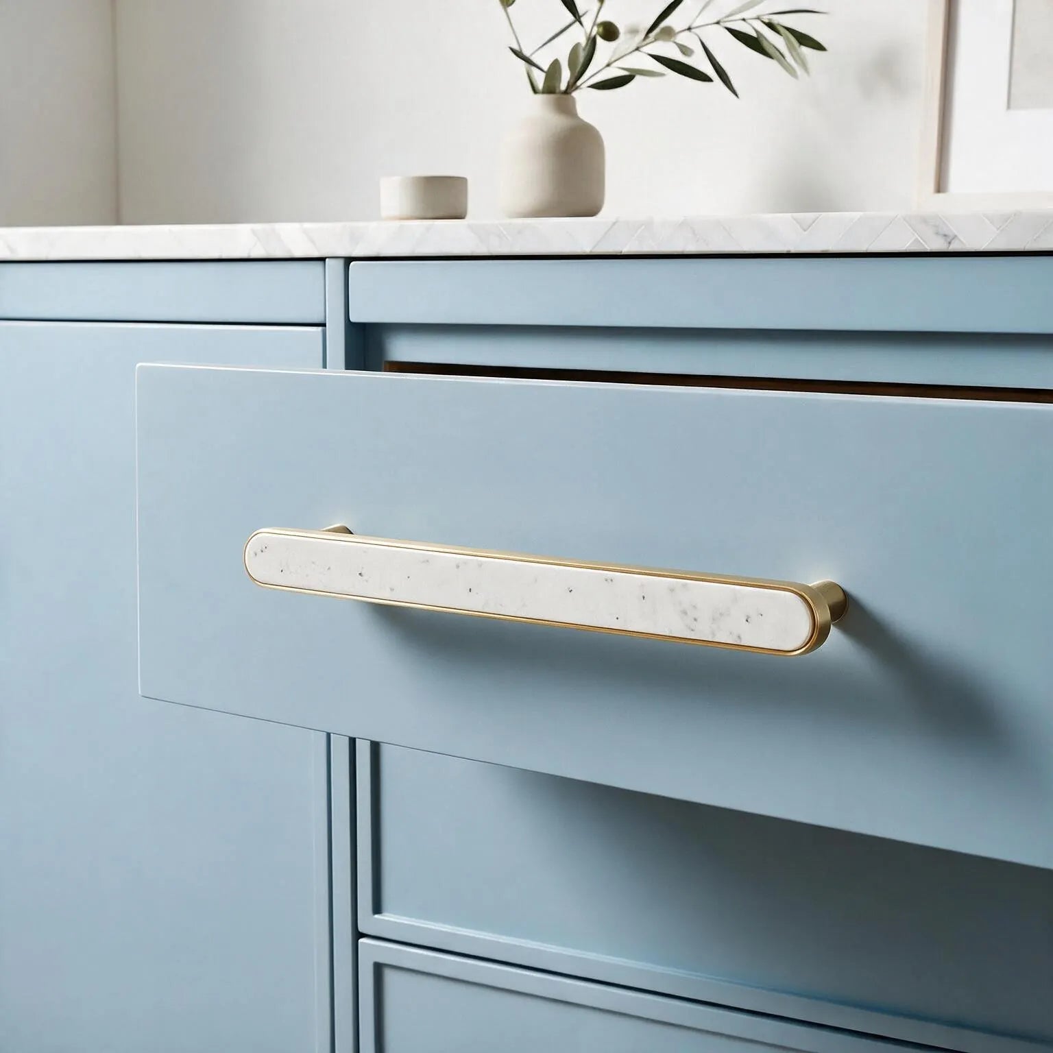 A close-up of a light blue cabinet drawer featuring a long horizontal bar handle with a white marble inlay and brushed gold-toned metal edges, set against a softly styled interior background.