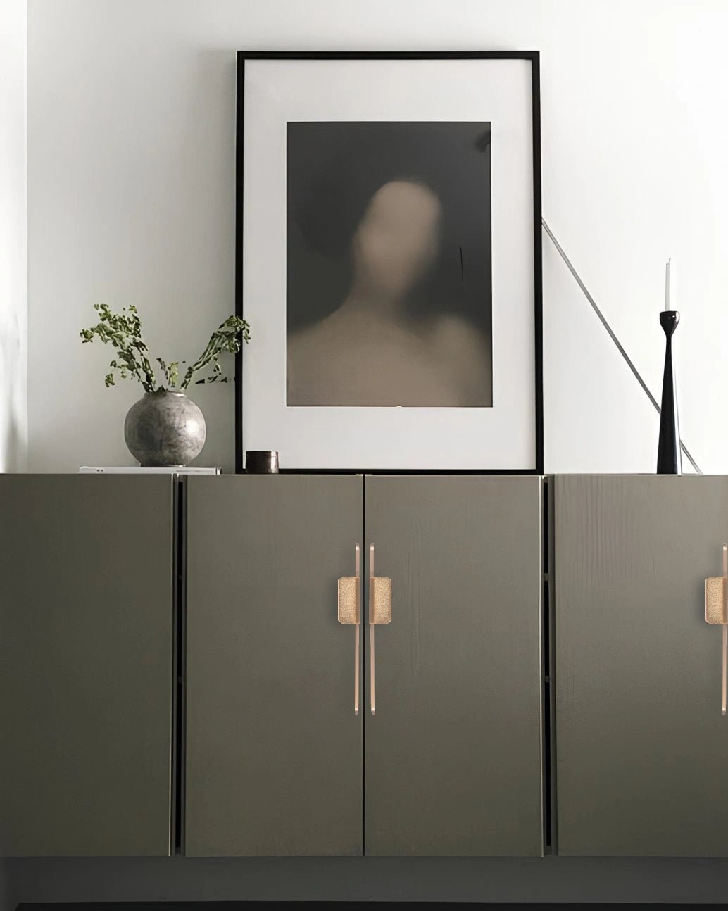 Modern interior with a dark green cabinet featuring sleek handles, framed abstract art, and a vase with greenery—by Mantara.