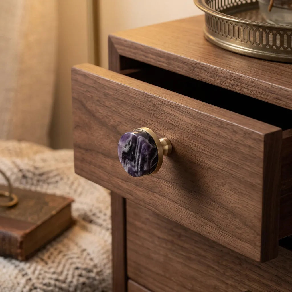 A close-up of a wooden nightstand or cabinet with a drawer pulled open, featuring a round purple marbled stone knob with a brass or gold-toned metal base. The warm wood grain is prominent, and a cozy home interior setting is visible in the softly lit background.