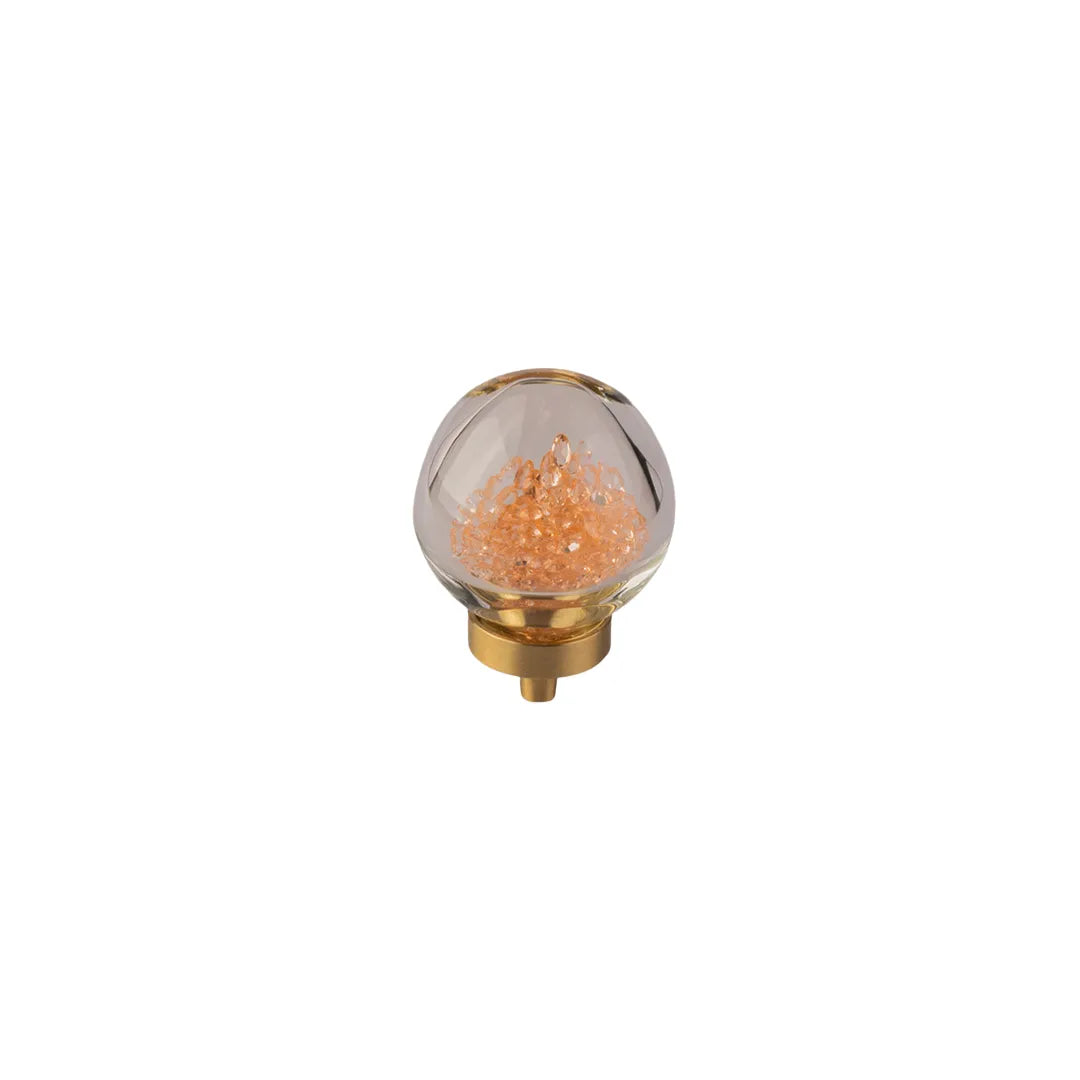 A clear glass teardrop-shaped decorative light or ornament with a gold-toned metal base and internal warm orange glitter or crystal-like fill, centered on a plain white background.