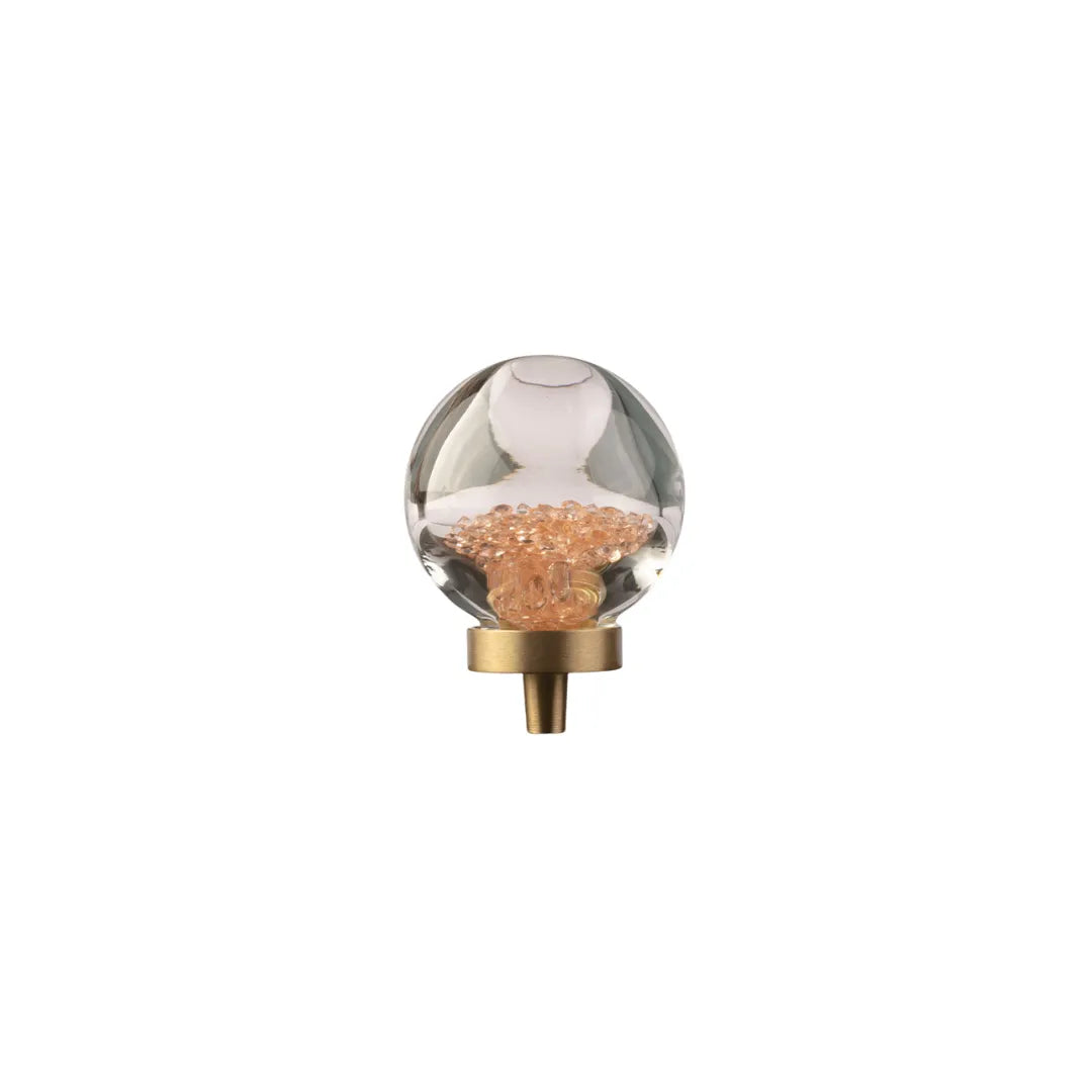 A modern wall sconce featuring a clear glass globe bulb filled with peach-colored decorative chips, mounted on a brushed brass circular ring with a short brass post.