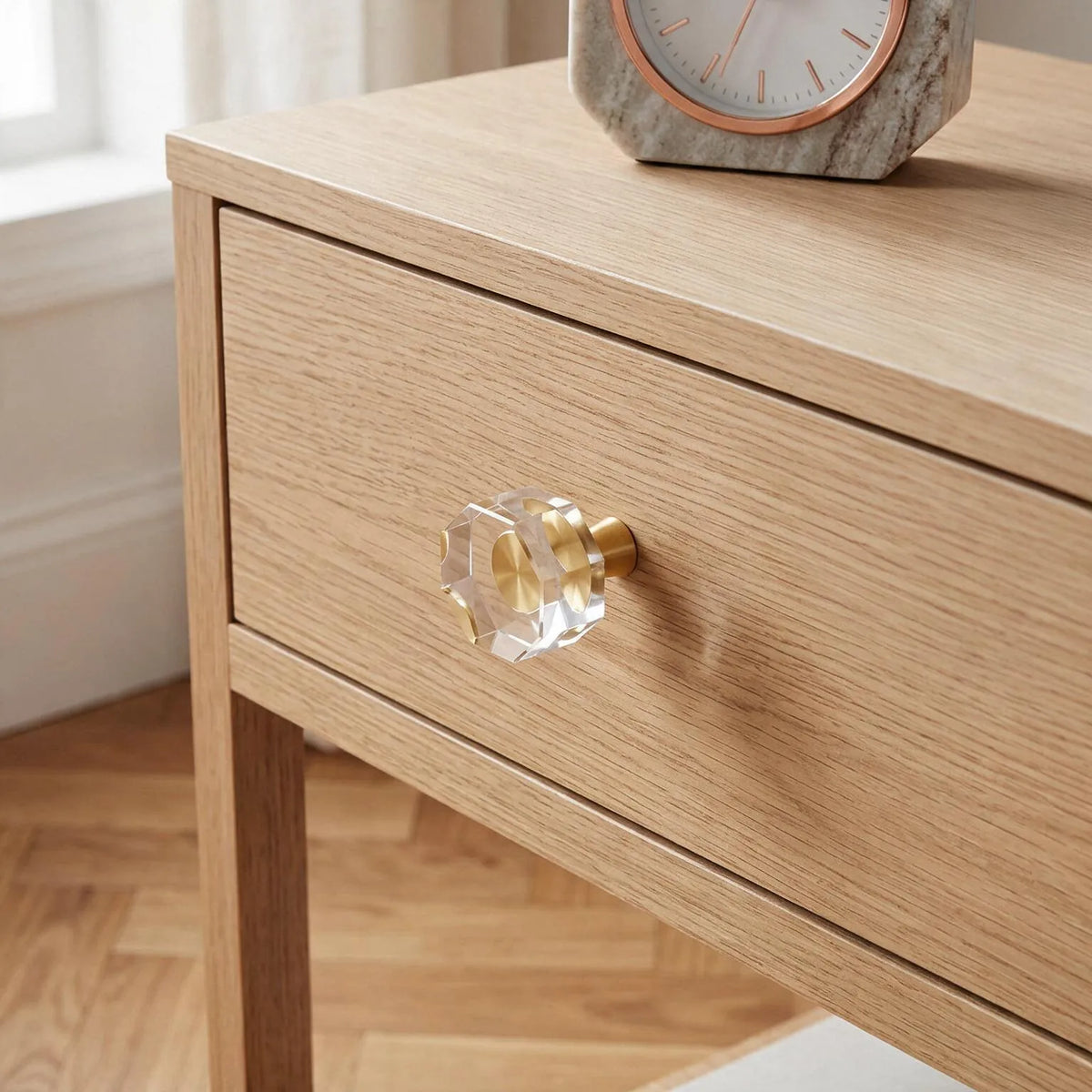 A close-up of a light oak wood nightstand with a clear faceted crystal drawer knob and a brass pull, shown in a bright room with a marble clock on top.