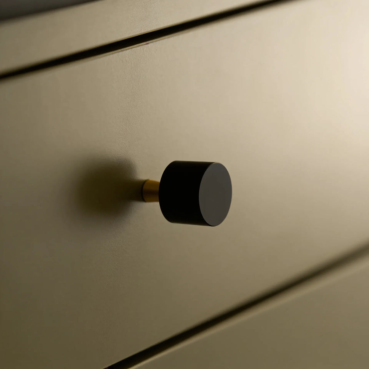 A close-up of a modern cylindrical drawer knob with a matte black top and a brass-toned base, mounted on a warm beige wood drawer front.