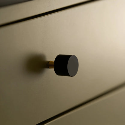 A close-up of a modern cylindrical drawer knob with a matte black top and a brass-toned base, mounted on a warm beige wood drawer front.