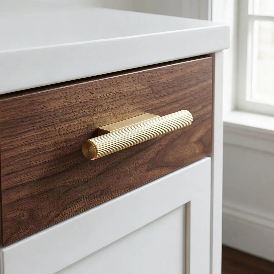 A close-up of a modern drawer featuring a dark brown wood grain front, a ribbed gold metal handle, and a white cabinet frame, with a softly blurred window in the background.