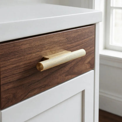 A close-up of a modern drawer featuring a dark brown wood grain front, a ribbed gold metal handle, and a white cabinet frame, with a softly blurred window in the background.