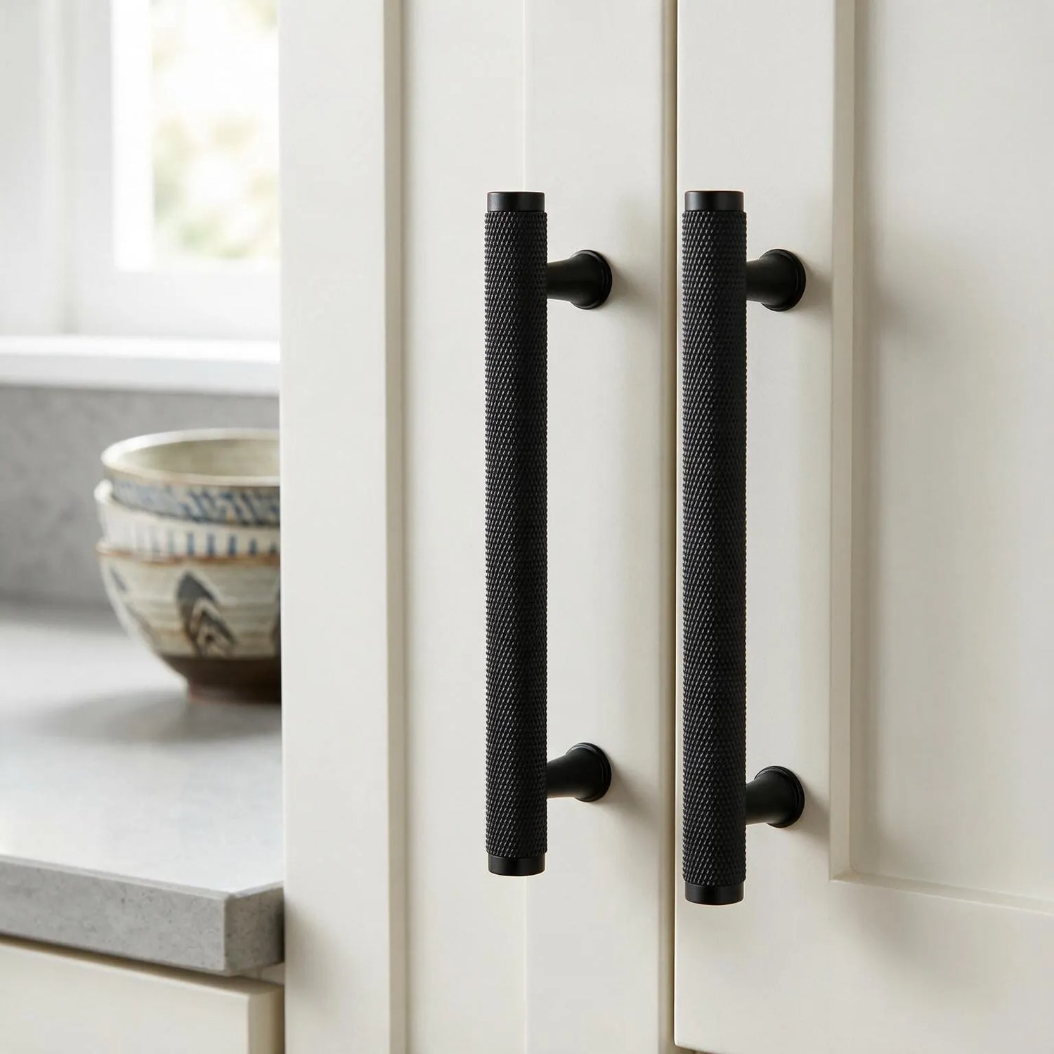 Two long black textured cabinet handles mounted on white kitchen doors, with a countertop and bowls visible in the softly lit background.