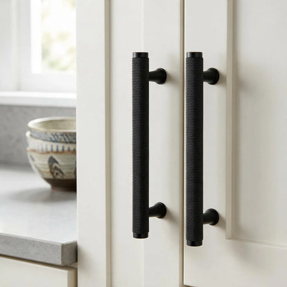 Two long black textured cabinet handles mounted on white kitchen doors, with a countertop and bowls visible in the softly lit background.