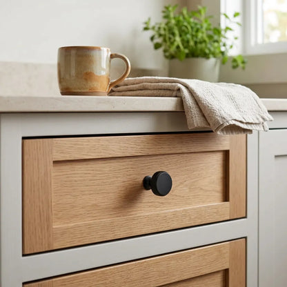 A close-up of a kitchen cabinet with light oak wood drawers and a matte black round knob, topped with a beige countertop, a ceramic mug, and a folded cloth, with a potted plant in the background.