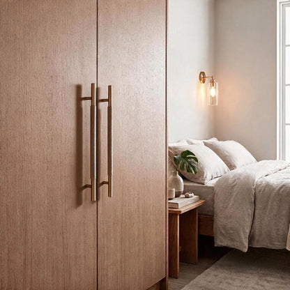 A pair of tall wooden wardrobe doors with long, brushed brass double handles, shown in a modern bedroom setting next to a neatly made bed.