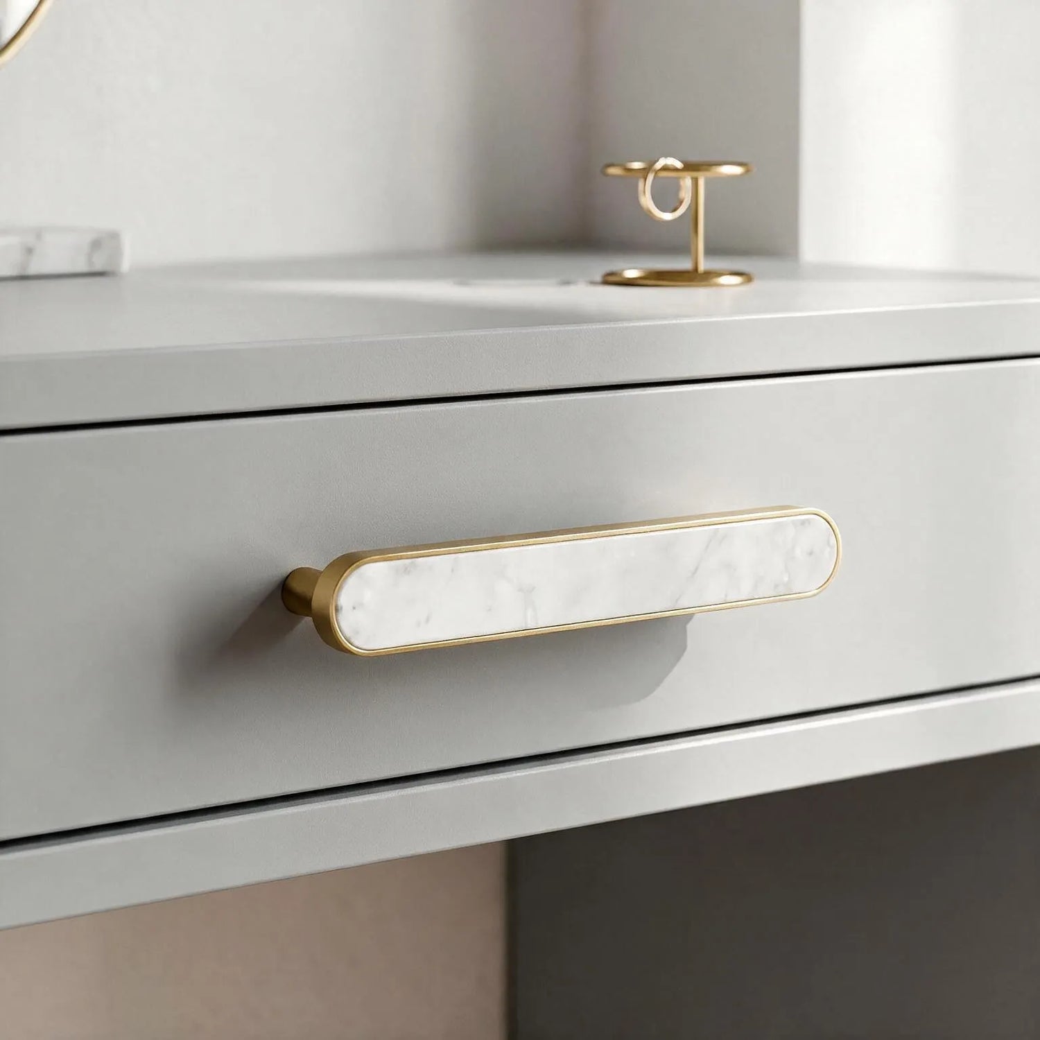 A close-up of a modern drawer featuring a long horizontal bar handle with a white inlay and brushed gold frame, set on a matching light gray vanity or desk with a small gold ring holder on top.