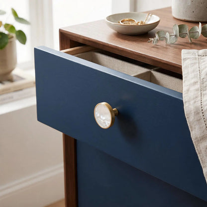 A close-up of a navy blue drawer on a wooden nightstand with a round brass knob, shown in a styled home setting with a bowl and eucalyptus leaves on top.