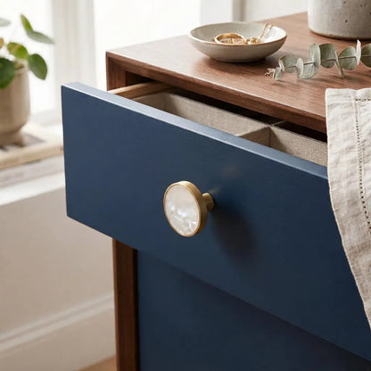 A close-up of a navy blue drawer with a round mother-of-pearl knob and a brushed gold frame, set on a wooden nightstand in a bright room.