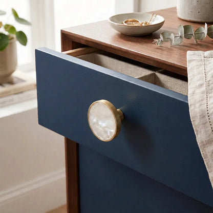 A close-up of a navy blue wooden drawer with an open top, featuring a round mother-of-pearl-style knob with a gold-toned frame. 