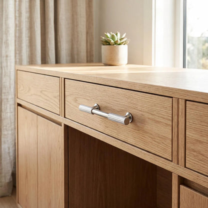 A light oak wood writing desk with a silver drawer pull handle, shown in a bright room next to a window with beige curtains and a small potted succulent on top.