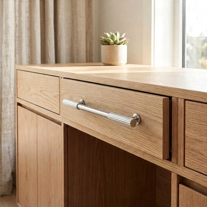A light oak wood writing desk with a silver drawer pull handle, shown in a bright room next to a window with beige curtains and a small potted succulent on top.