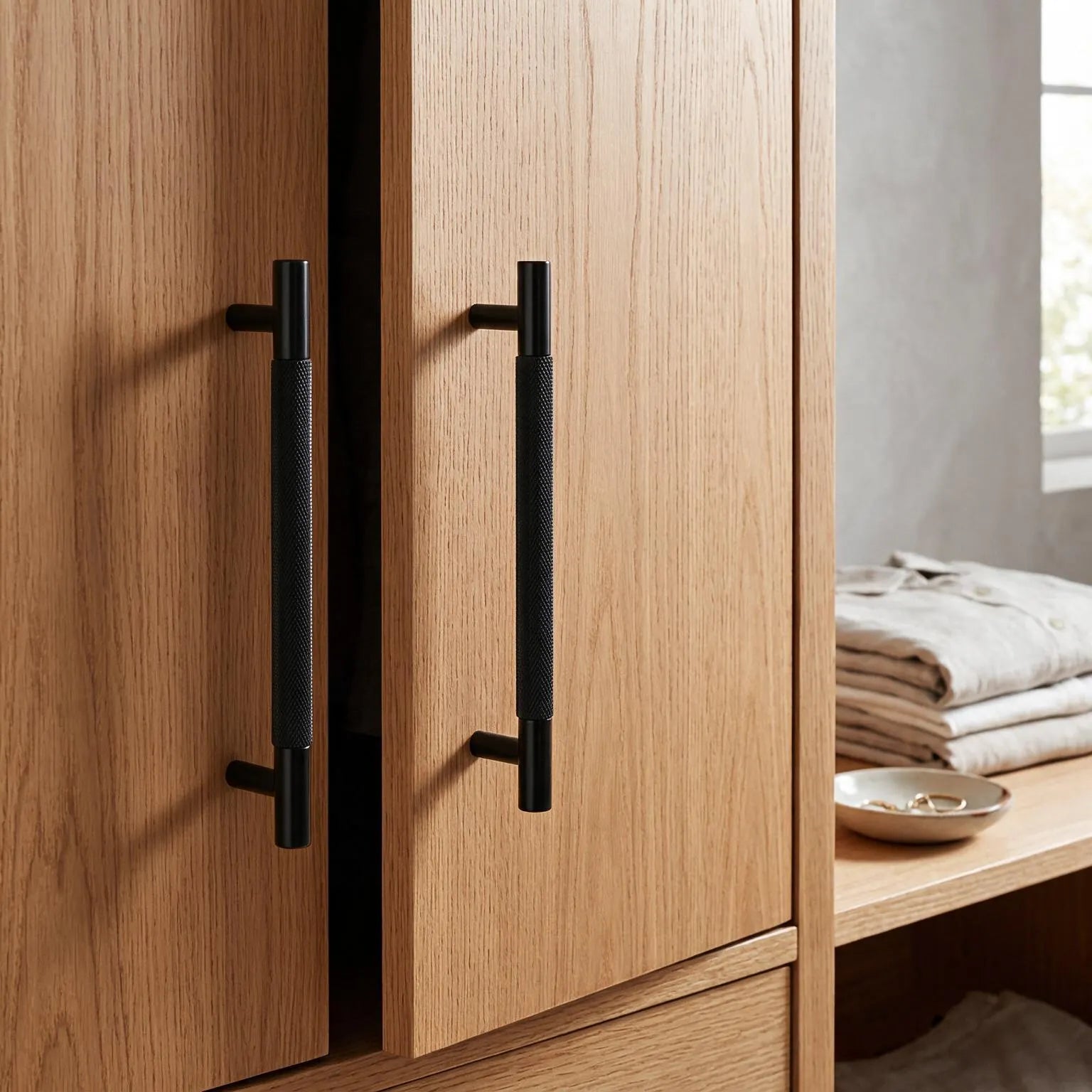 Two modern matte black bar handles with a textured grip finish mounted on light oak wood cabinet doors, shown in a styled interior setting with folded linens on a shelf.