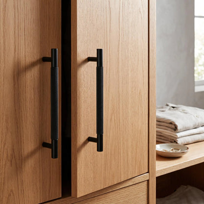 Two modern matte black bar handles with a textured grip finish mounted on light oak wood cabinet doors, shown in a styled interior setting with folded linens on a shelf.