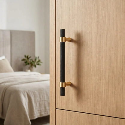 A modern bedroom scene showing a light wood wardrobe door with a long vertical black handle featuring brushed gold end caps, with a softly blurred bed in the background.