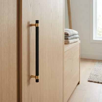 A modern long bar handle with a matte black center and brushed brass end caps, mounted on a light oak wood cabinet door in a bright, minimalist room.