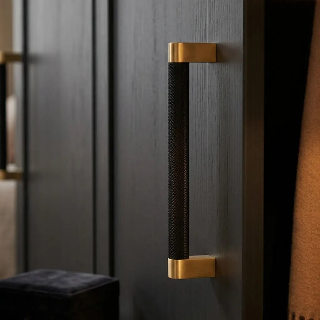 A modern vertical cabinet handle with a textured black center and brushed gold end caps, mounted on a dark wood door.