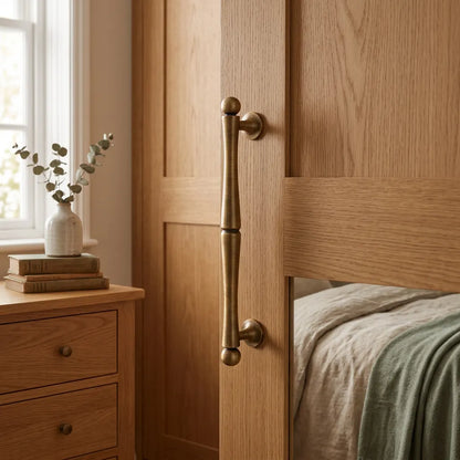 A close-up of a bedroom interior showing a light oak wood wardrobe door with a long, vertical, antique brass pull handle. The door is slightly open, revealing a bed with neutral-toned bedding and a green throw blanket.