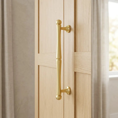 A long, vertical brass door handle with decorative ball finials at the top and bottom, mounted on light oak wood panel doors.