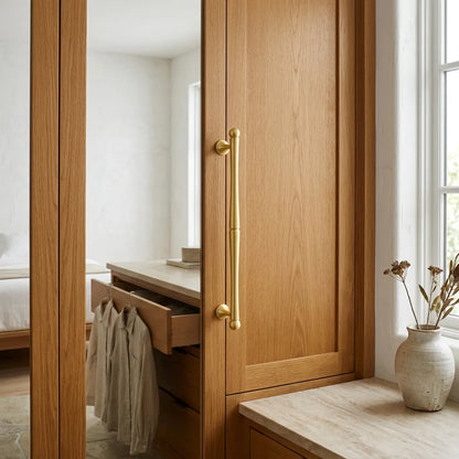 A close-up of a warm oak wood cabinet door with a prominent vertical brass handle, set in a bright, modern room with a mirror, open drawer, and a small vase on a windowsill.