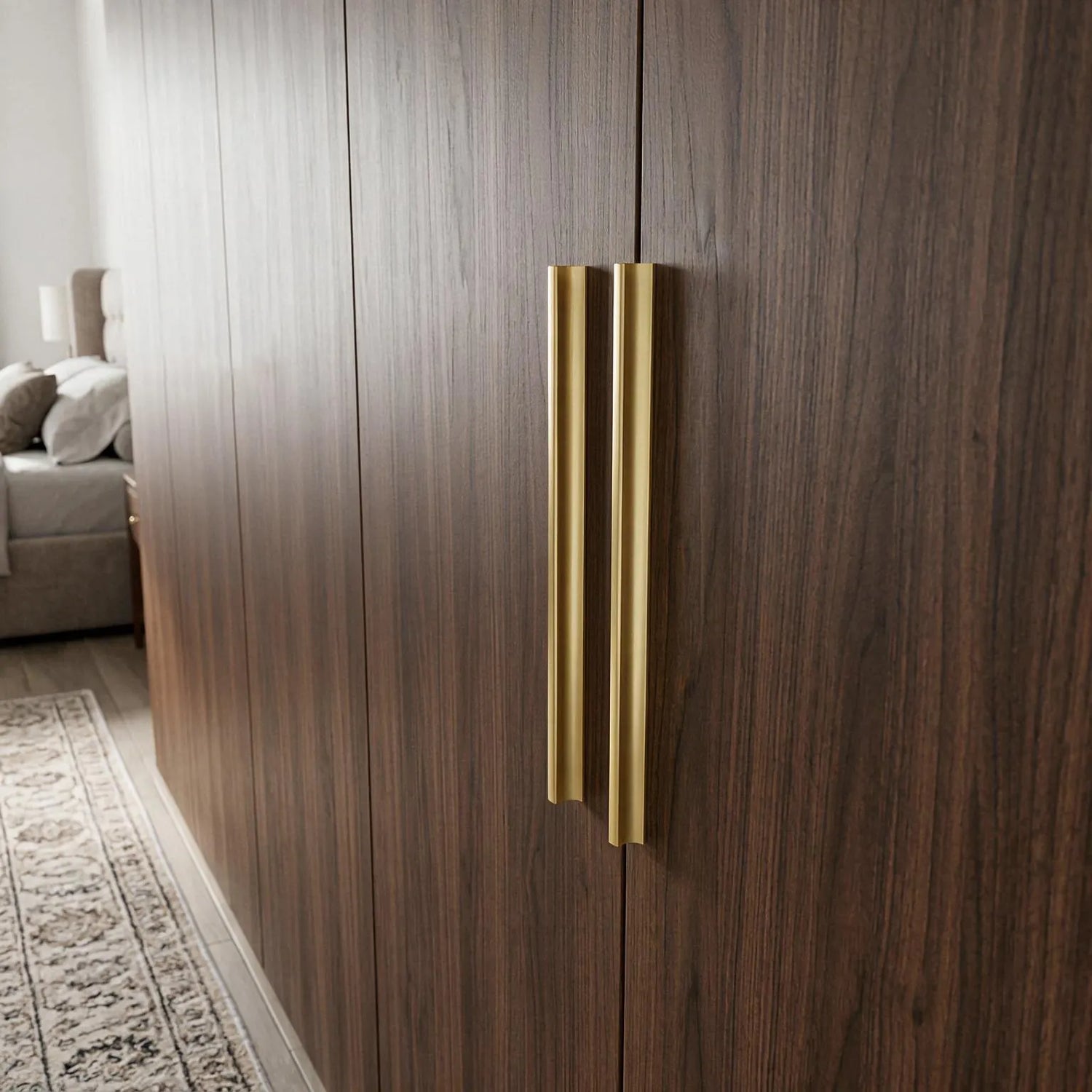 A pair of modern, slim vertical brass door handles mounted on dark wood grain doors in a bedroom setting.