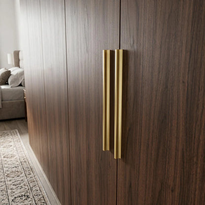 A pair of modern, slim vertical brass door handles mounted on dark wood grain doors in a bedroom setting.