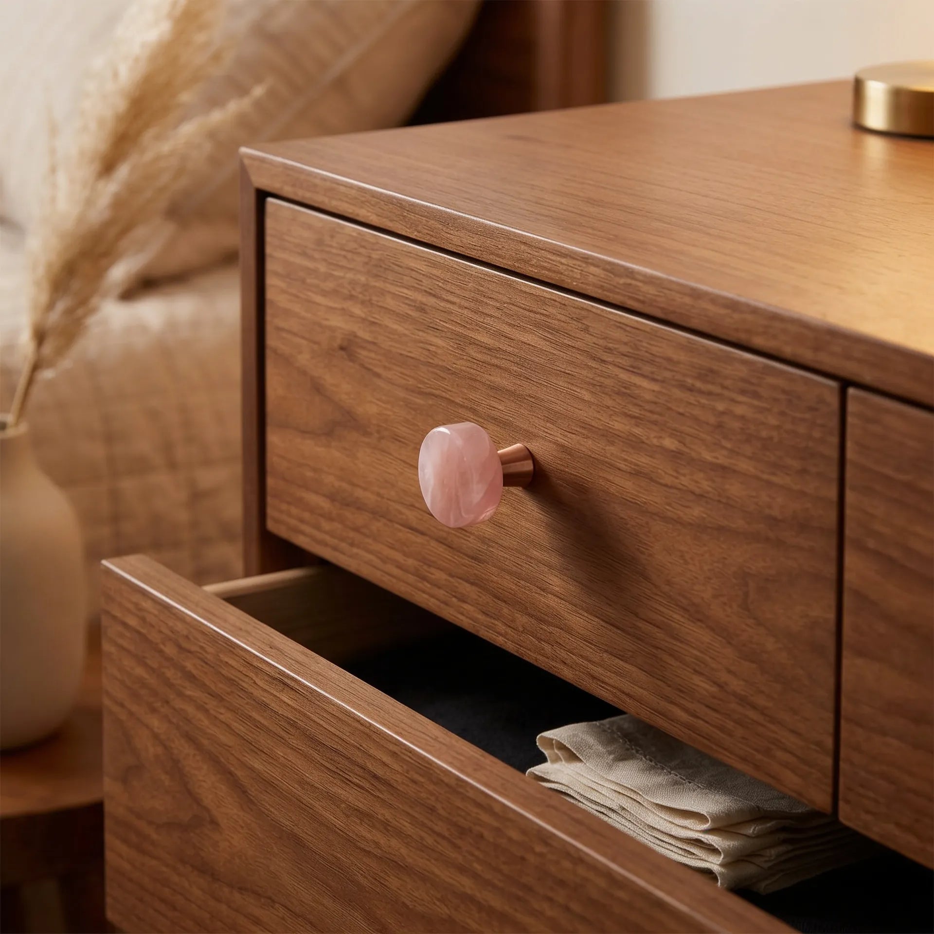 A close-up of a wooden nightstand or cabinet with a warm medium-brown wood grain finish, featuring a round pink stone drawer knob and an open lower drawer holding folded fabric.