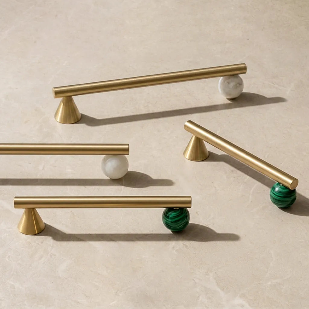 Four modern cabinet pulls with brushed gold bars and cone-shaped bases, shown on a light beige stone surface. Three pulls have white marble ball accents, and one has a green malachite ball accent.