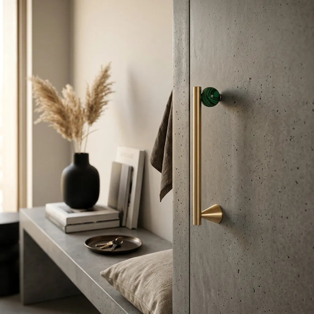 A modern interior scene showing a tall matte brass door handle with a round green knob, mounted on a speckled gray concrete door next to a matching concrete bench with decorative books and a vase.