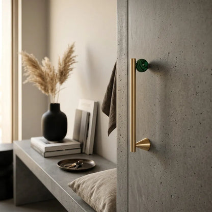 A modern interior scene showing a tall matte brass door handle with a round green knob, mounted on a speckled gray concrete door next to a matching concrete bench with decorative books and a vase.