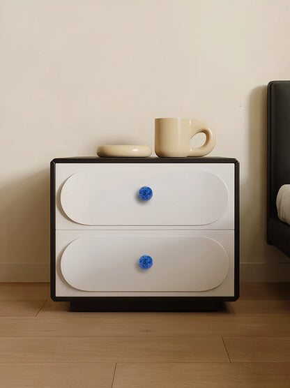 White nightstand with blue knobs, topped with a cup and saucer, placed on a wooden floor, styled by Mantara.