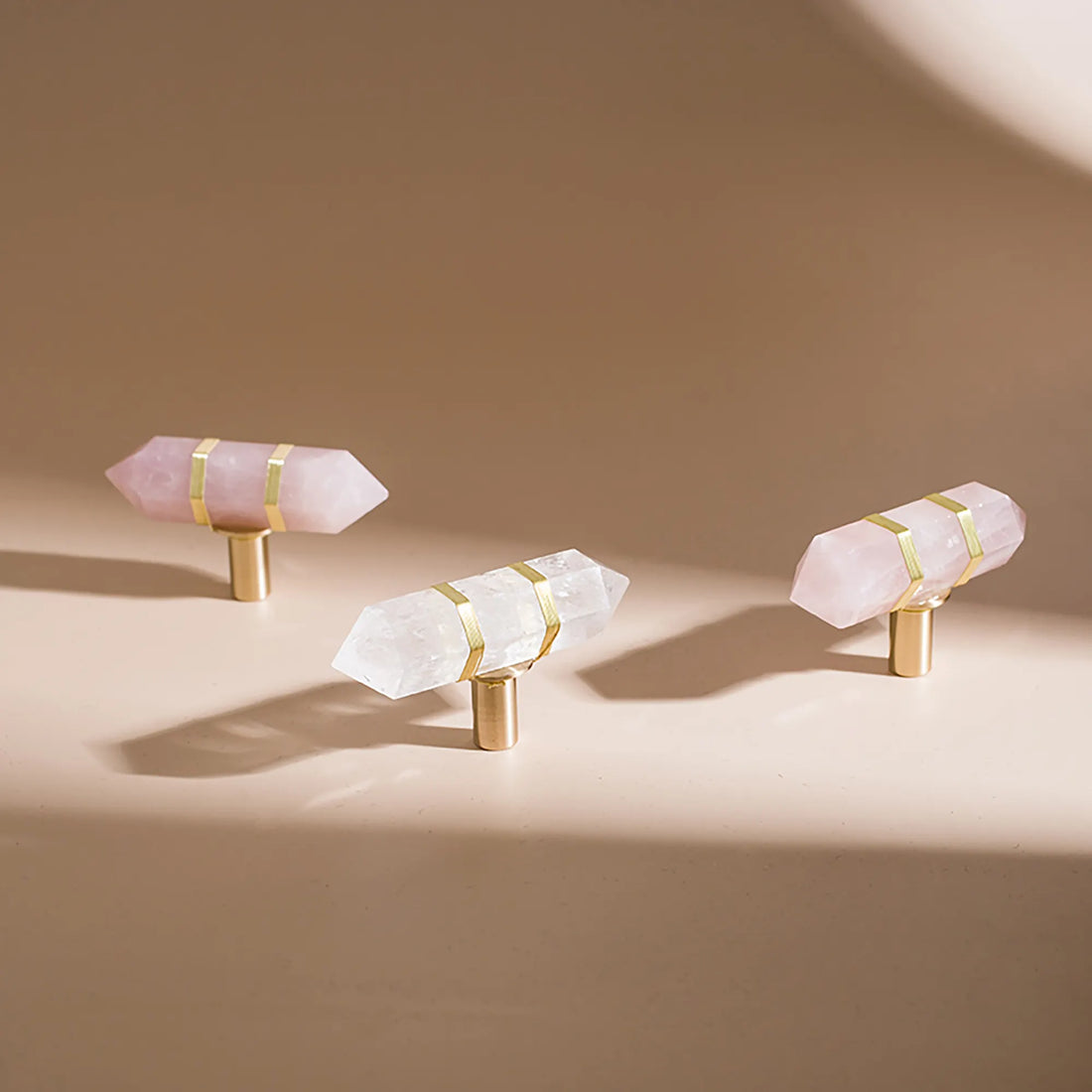 Three crystal point cabinet knobs with gold bands and gold cylindrical bases, displayed on a warm beige surface with soft shadows.