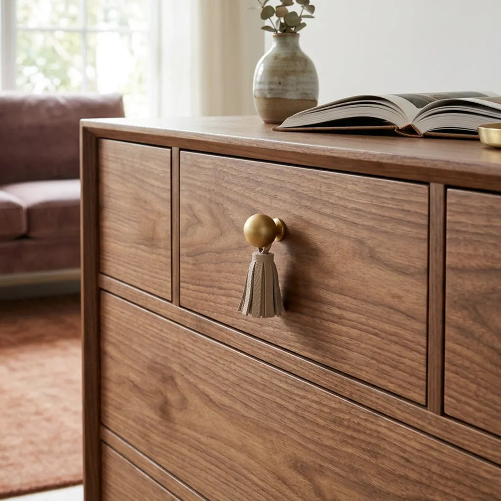 A close-up of a wooden dresser with a warm medium-brown finish, featuring a gold-toned tassel pull on the front drawer. 